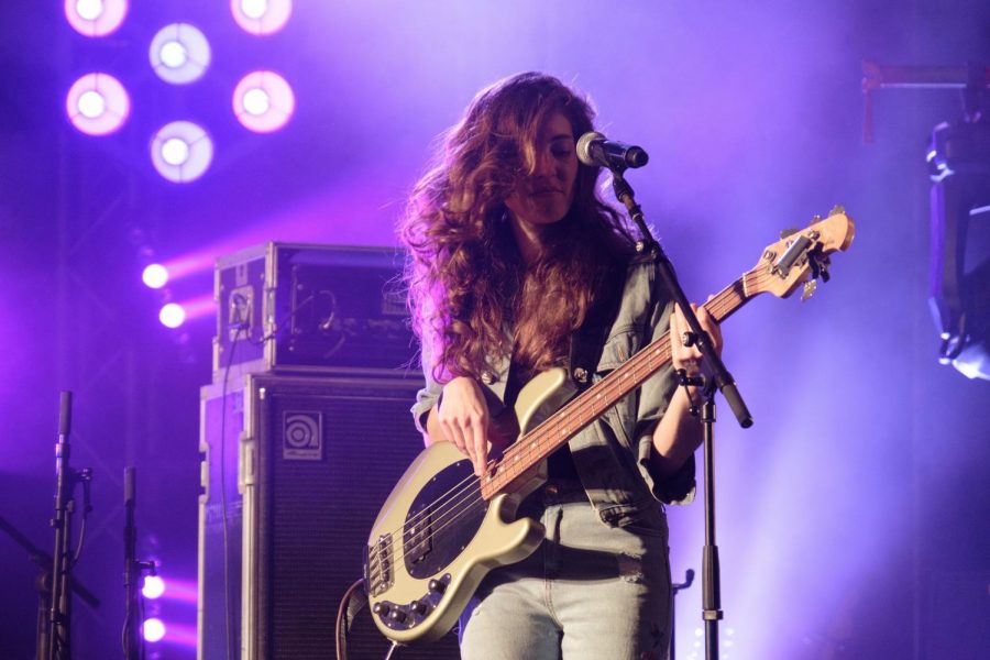 A brown-haired woman is playing the bass guitar on a stage. There are purple lights surrounding her and stage equipment.