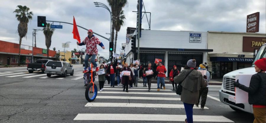 One LAUSD staff member balances on his unicycle on the streets of Van Nuys. Behind him, a crowd of people hold signs that read, “PAY US! RESPECT US!”.