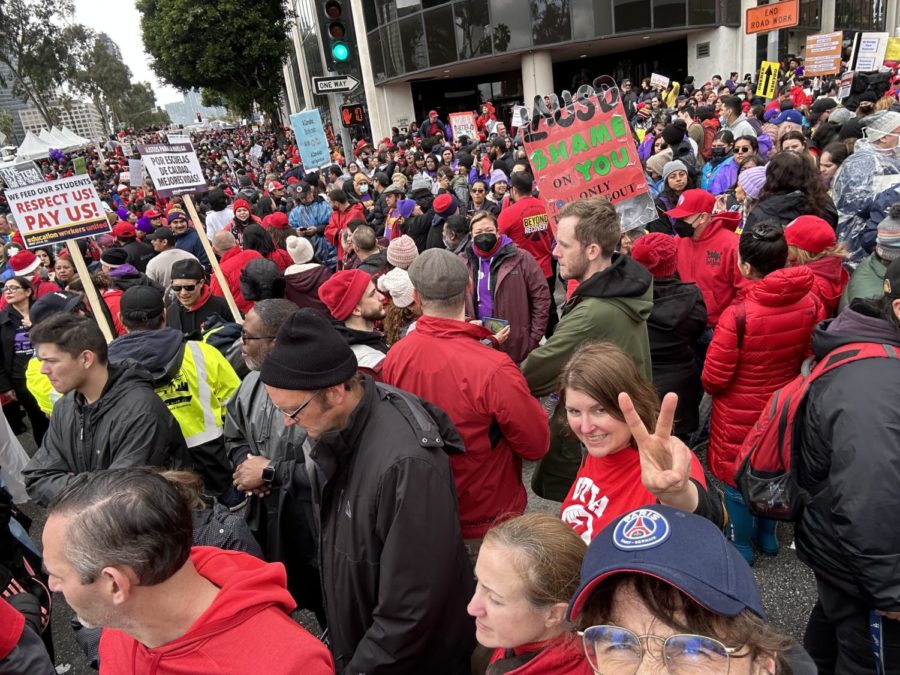 Hundreds of LAUSD staff members make a sea of red while huddling together in the rainy weather. A faculty member happily holds up a peace sign.