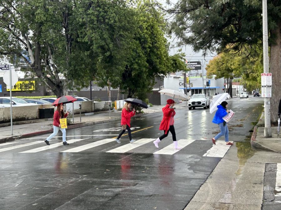 LAUSD staff members carry umbrellas while quickly running across the street while dressed in red.  